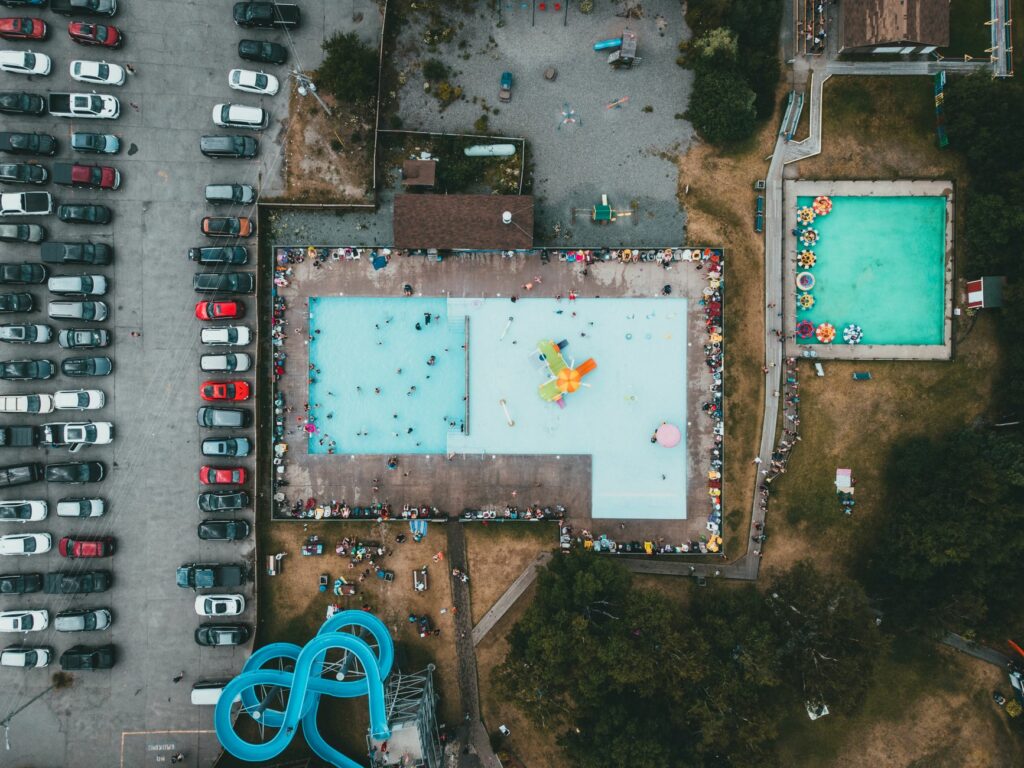 Aerial view showcasing a busy swimming pool surrounded by cars and parking area, emphasizing summer fun.